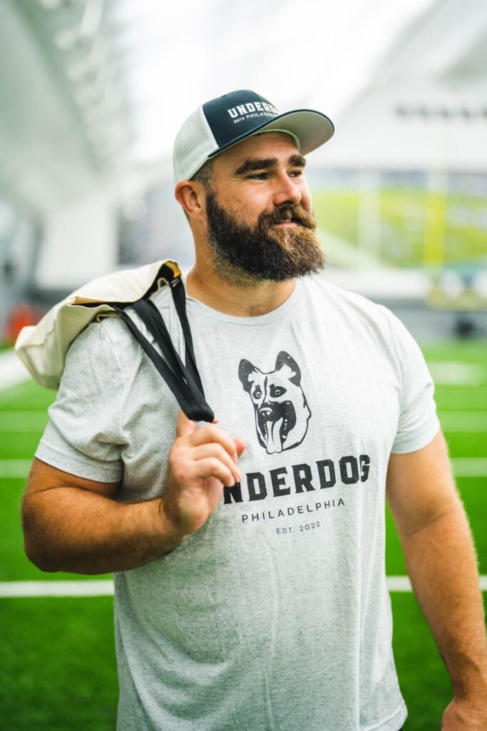 Jason Kelce looking right off camera modeling the Underdog Tee and Underdog Tote from the Underdog Apparel line