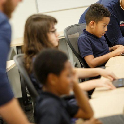 Three students on computers at a desk learning from two teachers