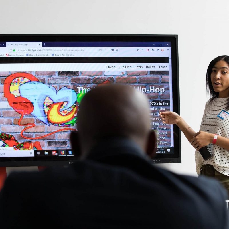 Point of view looking at the back of a man's head sitting in front of the camera with a teacher and interactive whiteboard behind him